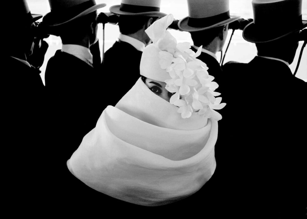 Probablement une des plus élégantes photographies de Frank Horvat, réalisée pour Givenchy et le Journal Jardins des Modes. Le modèle féminin porte un chapeau et est drapé dans un large foulard tandis qu'à l'arrière plan des messieurs en hauts de forme regarde une course de cheveaux.